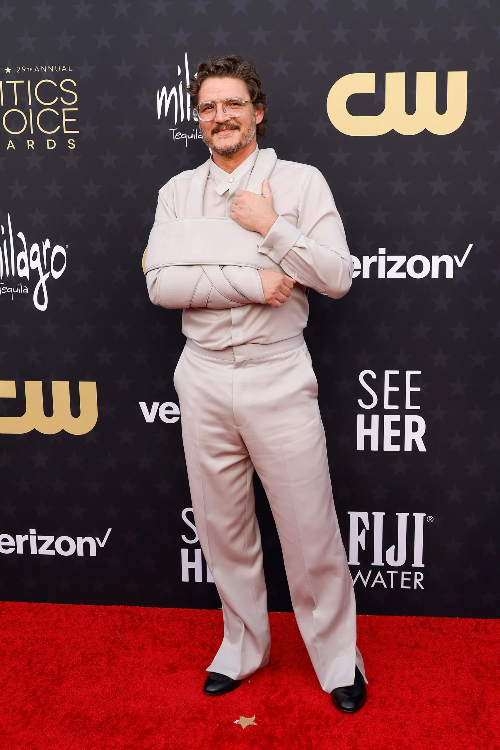 As at last week’s Golden Globes, Pedro Pascal incorporated his arm sling into his look — this time, a gray-beige outfit paired with clear spectacles and black shoes.
